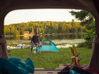 A mother and daughter playing soccer next to a tent by a lake.