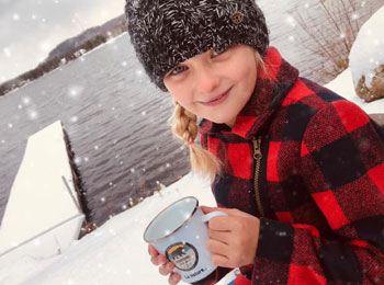 Young girl with a hat and plaid shirt by the water in the winter holding a mug