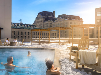 A few people swimming in an outdoor pool; there’s snow on the ground and historic buildings on the horizon.