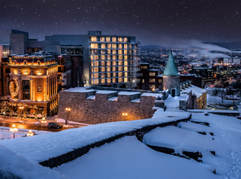 The hotel’s modern building amidst historic buildings under snow; the sky is starry and the city is illuminated in the distance.